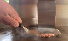 A flat metal spatula pressing a burger.